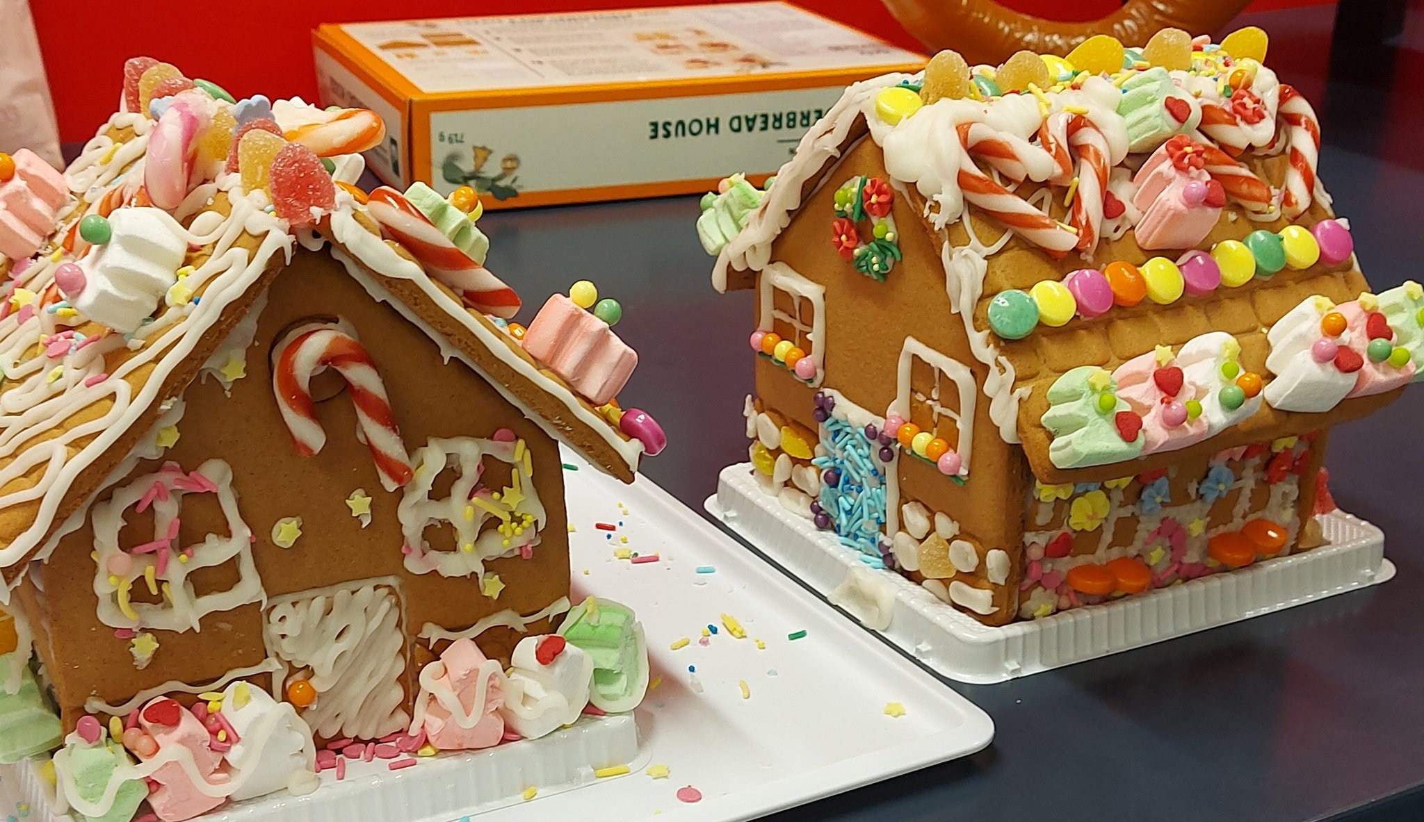 two gingerbread houses decorated with icing, candy, marshmallows, and sprinkles are displayed on white plates. a gingerbread house kit box is in the background.