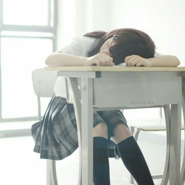 a schoolgirl in a white shirt, plaid skirt, and knee socks is sleeping with her head on a desk in a classroom. she is sitting in a chair, and the desk is visible in the foreground. the classroom appears bright, with a large window in the background.