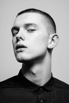 a black and white portrait of a young man with a buzz cut, staring intensely at the camera. he has a serious expression and a piercing in his left earlobe. he is wearing a black buttondown shirt. the photo is taken in a studio with soft lighting.