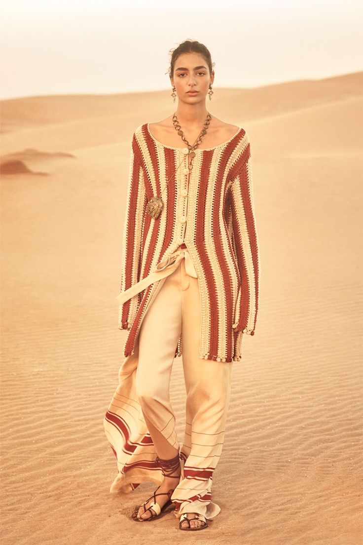 full shot of a fashion model, a woman with light tan skin, wearing a longsleeved, striped crochet cardigan in shades of red and cream, paired with wideleg, creamcolored pants that feature subtle red stripes along the hems.  she accessorizes with a long, ornate necklace, dangling earrings, and simple brown leather sandals.  the model is standing in a sandy desert landscape, the texture of the sand visible in the background. the overall aesthetic is bohemian and chic. the lighting is warm and natural, suggestive of a fashion photoshoot.