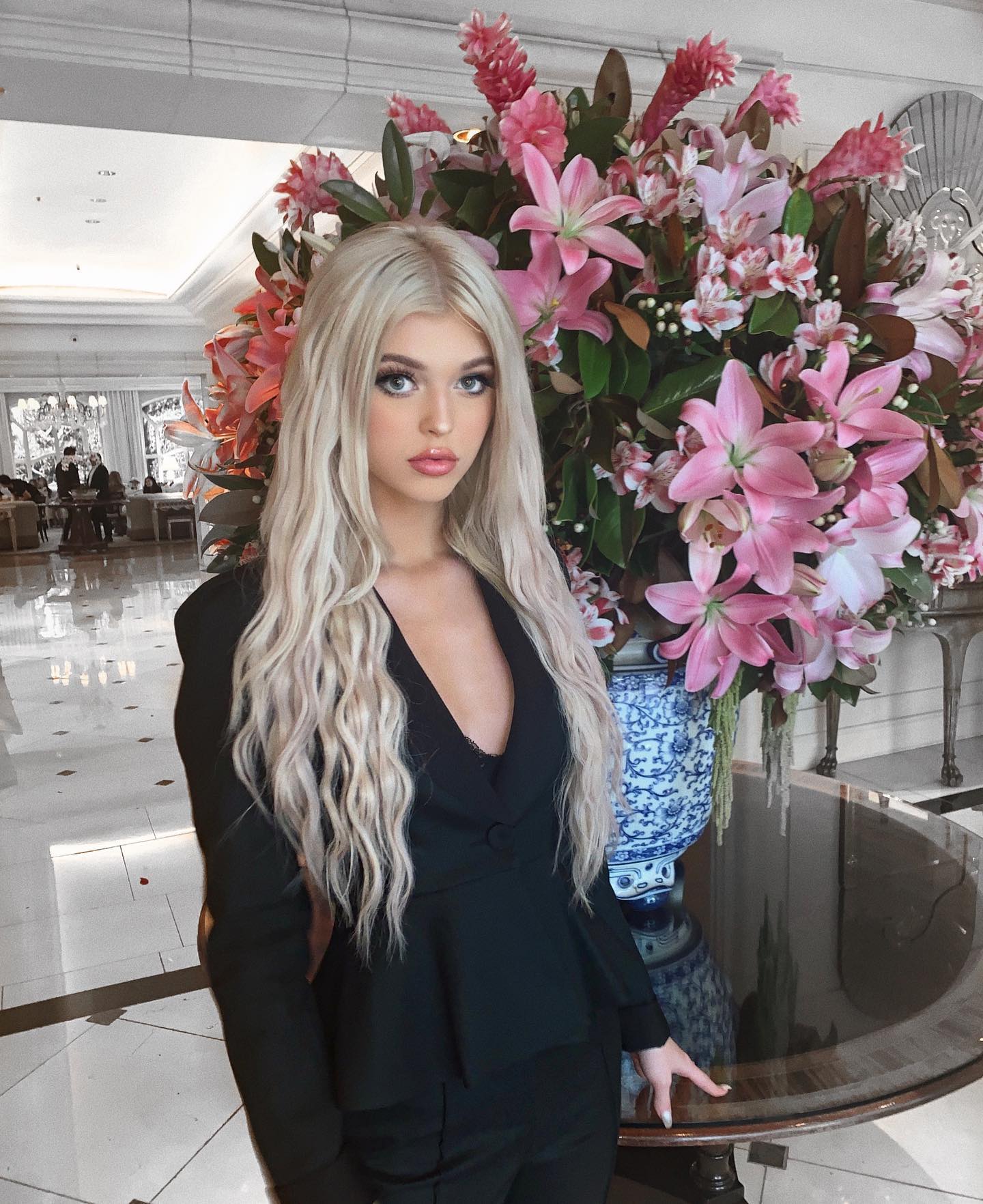 a woman with long blonde hair is standing in a hotel lobby next to a large arrangement of pink lilies in a blue and white vase on a round glass table. she is wearing a black suit.