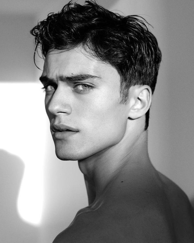 the image is a black and white, closeup portrait of a young, attractive male model.  he has dark, short, wavy hair styled back. his expression is serious and intense.  the lighting is dramatic, creating strong contrasts and shadows on his face and shoulders. the overall aesthetic is stylish and highfashion. the background is blurred and out of focus, drawing attention to the model's face and upper body.  the image is cleanly composed and professionally shot.