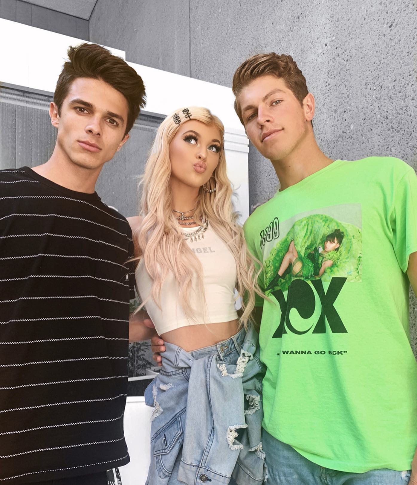 a waistup shot of three people standing close together. on the left is a man with dark hair wearing a black striped tshirt. in the middle is a woman with long blonde hair, wearing a white crop top with "angel" printed on it, a denim jacket around her waist, jewelry, and a decorated hair clip. she is pouting and looking up. on the right is a man with brown hair wearing a green tshirt with a graphic on it. the background is a gray wall and a bright white window.