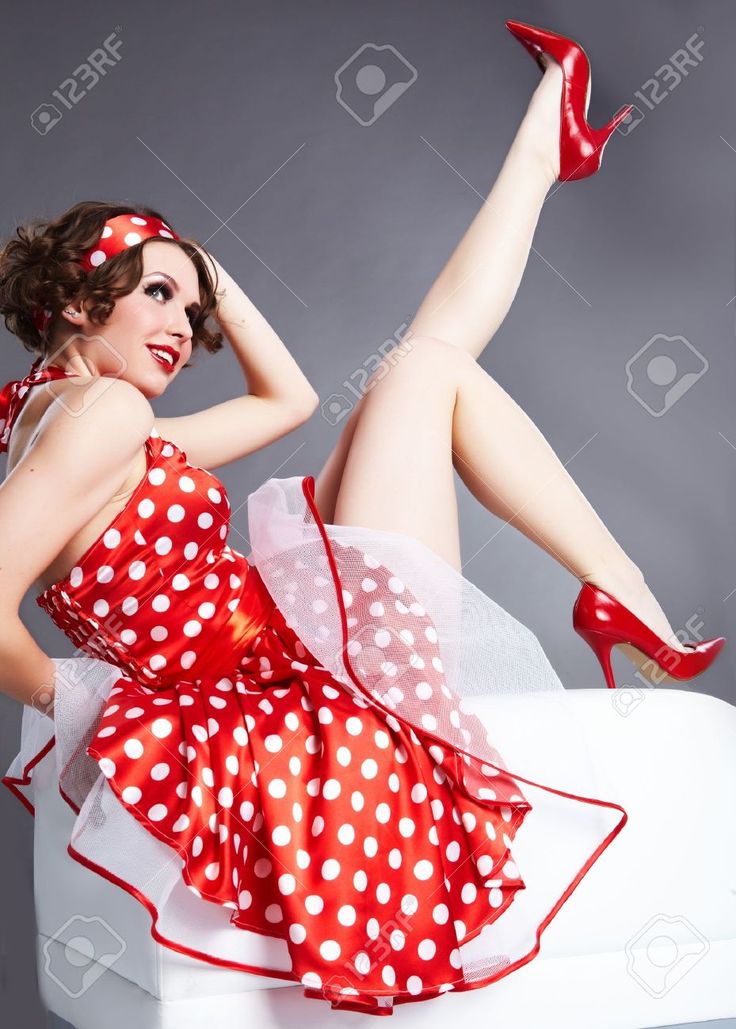 a captivating image of a pinup girl in a vibrant red polka dot dress.  she's sitting on a white surface, one leg elegantly raised, showcasing her long legs in red high heels. her retro hairstyle, headband, and makeup complete the classic pinup look. the gray background provides a neutral backdrop that accentuates the subject's red attire. the overall tone is glamorous and suggestive of vintage fashion and beauty.