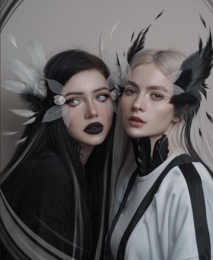 two women stand side by side looking at the camera. the woman on the left has long black hair with white streaks,  black clothing, and a white feather headband. she is wearing dark lipstick and dark eyeshadow.  the woman on the right has  long white hair and black feather headband,  wearing a white and black jacket. both women are wearing  decorative headbands with feathers. the background is a blurry image of a wall.