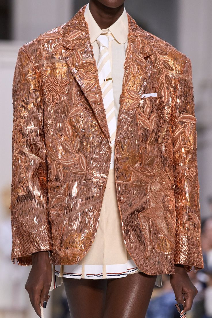 a fashion model is wearing a sparkling gold sequined blazer that has beautiful floral embroidery. she is also wearing a white skirt with a pleated hem.  the model is walking down a runway in what appears to be a fashion show.