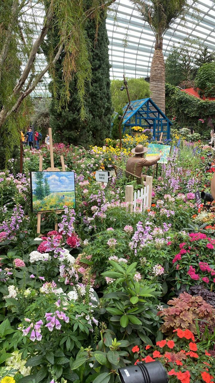 a vibrant and colorful garden scene is captured in the image.  numerous flowering plants in various shades of pink, purple, red, and white fill the foreground and middle ground.  there's a small blue greenhouse structure visible in the background.  a bronzecolored statue of an artist painting is situated amongst the flowers,  with an easel and a painted canvas depicting a similar landscape. people are visible in the background but distant and out of focus. the garden is enclosed within a large glassroofed structure that lets in natural light, creating a bright and cheerful atmosphere. the overall composition is a mix of natural beauty and artistic representation.