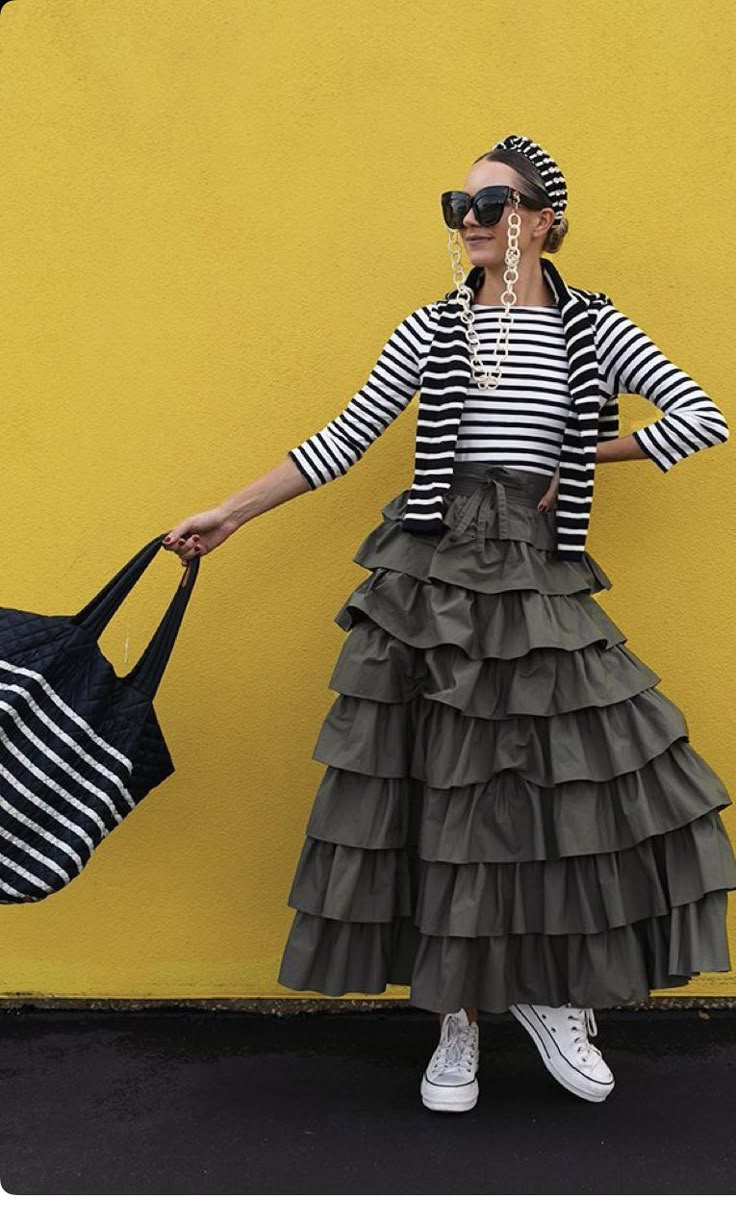 a woman stands in front of a bright yellow background, wearing a striped top and matching cardigan, paired with a voluminous, ruffled olive green skirt. she accessorizes with a black and white striped headband, oversized sunglasses held by a white chain, and white sneakers. she is holding a striped tote bag in her right hand.