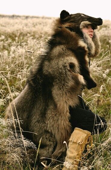 a person wearing a full fur coat and headdress in a grassy field. the headdress appears to be the head of a wolf or bear. the person is also wearing fringe boots.