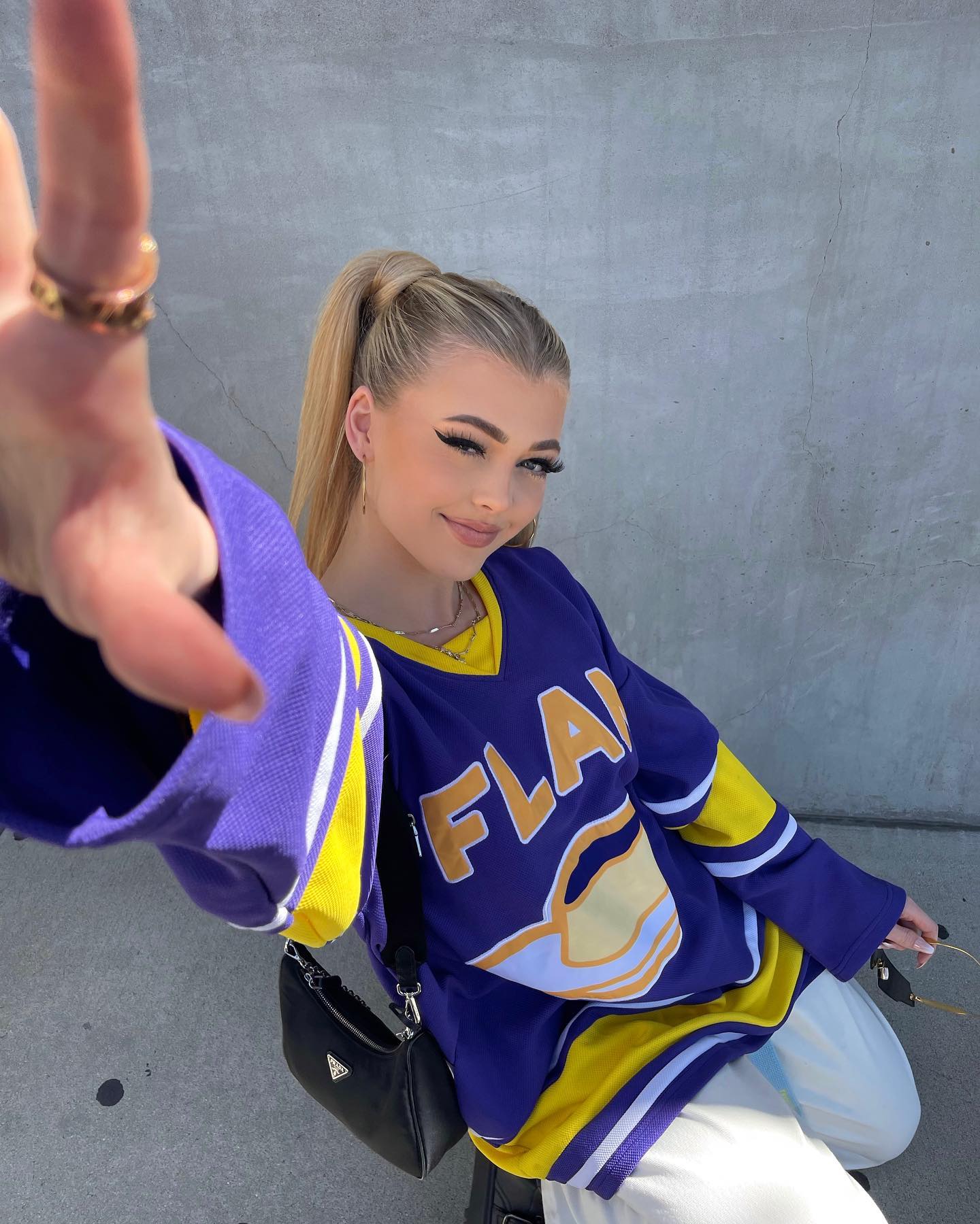 a woman with blonde hair in a high ponytail poses for a photo, wearing a blue and yellow hockey jersey and a black shoulder bag. she is smiling and holding her hand out towards the camera, showing off a ring. she is standing against a concrete wall.