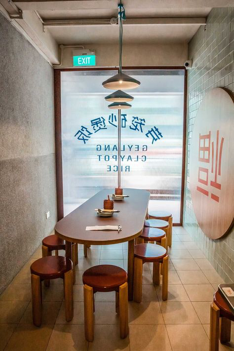 a clean and minimal restaurant interior with a wooden table and stools lined up on one side, a hanging lamp above the table and a window behind it. the window has a chinese sign and the text reads "rice clay pot geylang". the floor is tiled in light brown and the walls are beige. the overall atmosphere is cozy and inviting.