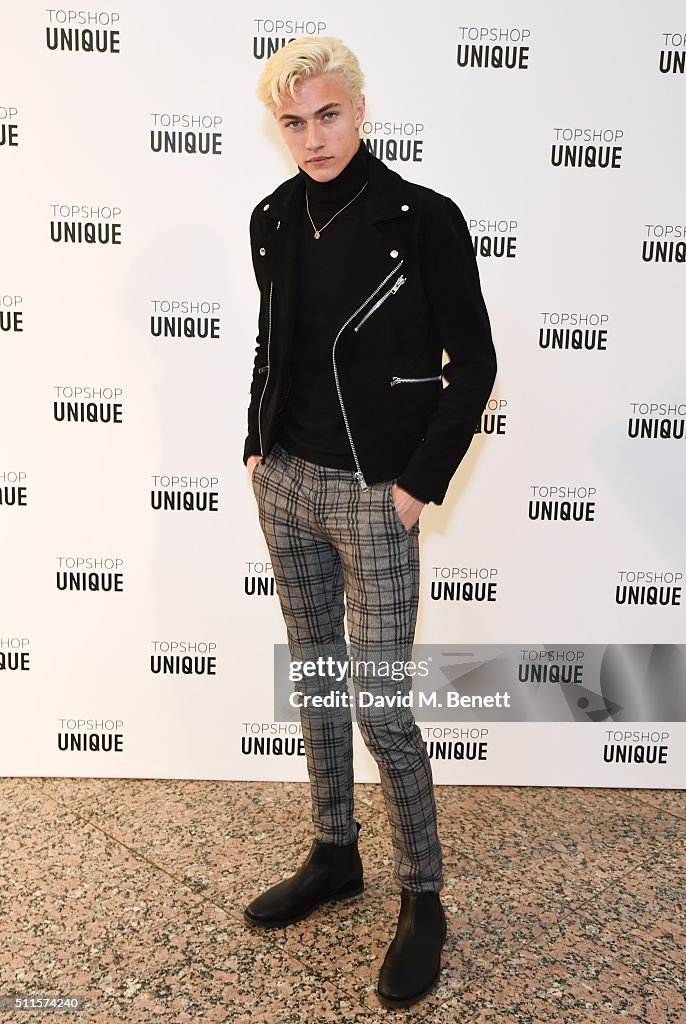 a young man with blonde hair is standing in front of a "topshop unique" branded backdrop. he is wearing a black turtleneck, a black jacket with silver zippers, plaid pants, and black chelsea boots. he is posing for the camera. the image also features a "getty images" watermark.