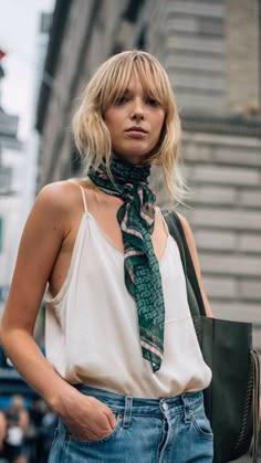a blonde woman in a white shirt, jeans, and a green patterned scarf smiles at the camera on a city street. she's also carrying a black tote bag.