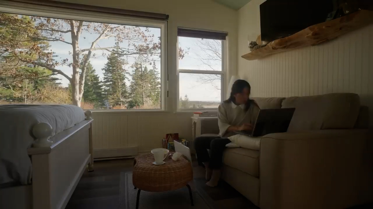 a woman sits on a couch in a bedroom, working on a laptop. she is positioned in front of two large windows that offer a scenic view of trees and the sea. the room is bright, illuminated by natural light streaming through the windows. a white coffee mug and a book rest on a small table beside her. the overall ambiance is one of peace and tranquility, suggestive of a comfortable workfromhome setting.