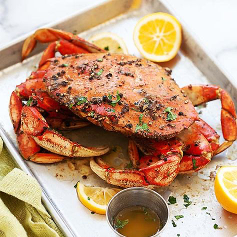 a closeup shot of a large, roasted dungeness crab sits on a metal baking sheet. the crab is generously seasoned with pepper and herbs, and garnished with fresh parsley.  lemon wedges are scattered around the crab, along with a small metal ramekin containing melted butter, likely for dipping.  the crab appears juicy and flavorful. the background features a partially visible light green linen napkin, enhancing the overall presentation of the delicious seafood dish.