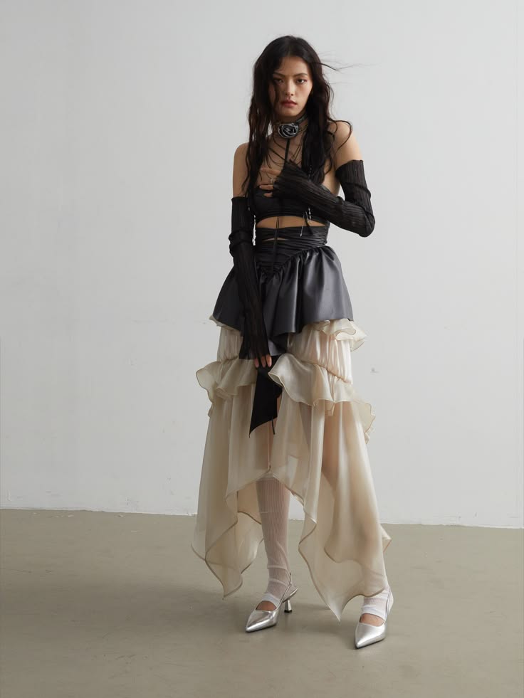 a woman model poses on a white background, wearing an allblack outfit with a black corset top and an asymmetrical, layered skirt with white ruffles. she has black gloves, silver shoes, and a silver choker necklace. her hair is straight and dark.  she appears to be on a runway or posing for a fashion shoot.