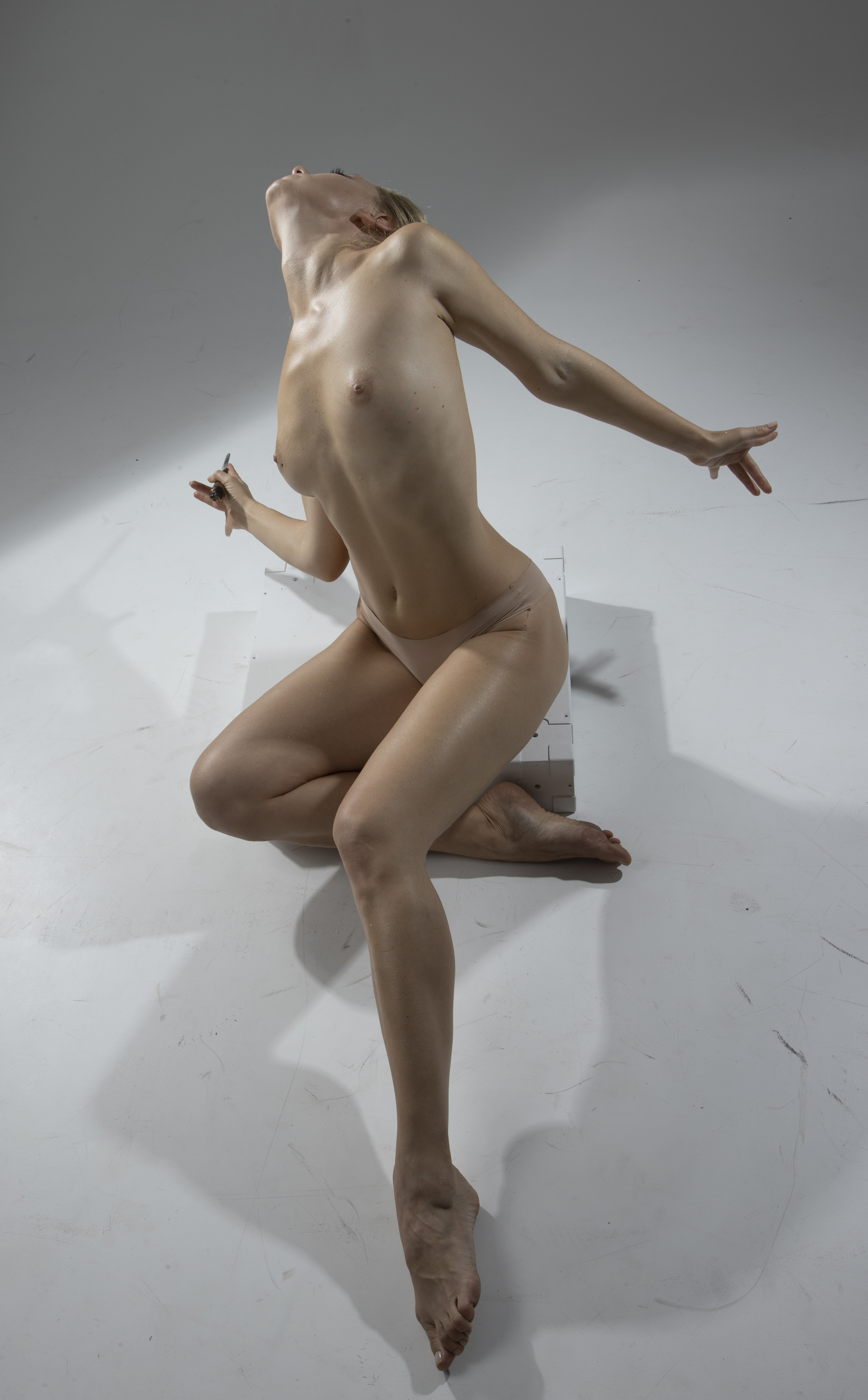 a nude woman is posing artistically in a studio setting. she is kneeling on a white, blocky object against a white background. her head is tilted back, and her arms are extended to the sides. her pose is expressive and highlights her bare torso and limbs. the lighting creates shadows on the background and her body.