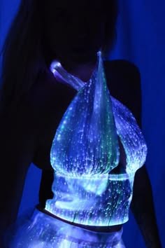a woman wears a futuristic top and skirt that glows brightly under a dark blue light. the outfit is made of fiber optic material that creates a dazzling display of light. the woman appears to be at a night club or rave, enjoying the vibrant atmosphere.