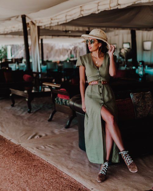 the image showcases a stylish woman in a flowing olivegreen maxi dress with a thighhigh slit.  she's wearing a beige safari hat, sunglasses, and brown ankle boots. a brown leather belt cinches her waist. the dress has a buttondown front.  she's accessorized with a delicate necklace. the background features an upscale outdoor setting, possibly a restaurant or lounge area within a tent, suggesting a luxurious travel or vacation destination, potentially in india (based on the style). the overall aesthetic is relaxed yet elegant, with a vintage or bohemian feel. the woman is sitting on a plush seat, appearing confident and comfortable.