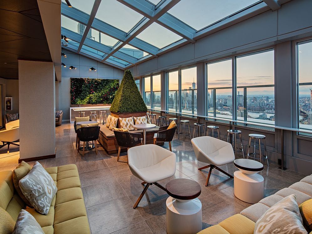an urban lounge space with modern chairs, couches, and tables, set on a rooftop with a panoramic view of the city skyline. the interior features a glass ceiling that allows natural light to fill the space. the lounge is decorated with green plants and has a sleek, contemporary design.