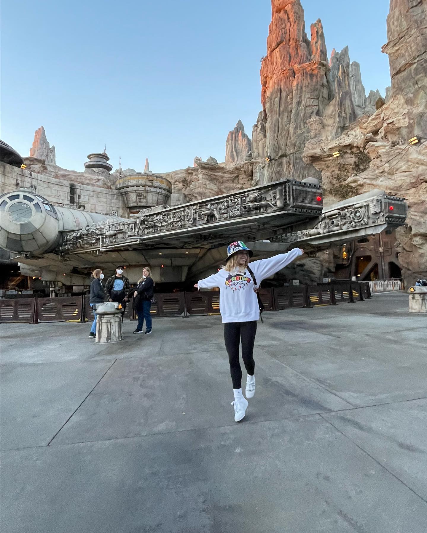 the image shows a young woman joyfully jumping in front of the millennium falcon ride at star wars: galaxy's edge in either disneyland or disney world.  she's wearing a white sweatshirt with a star warsthemed design, a bucket hat, and white sneakers. the background features the iconic millennium falcon, partially nestled within the rocky, desertlike landscape of batuu.  other people are visible in the distance, standing near the ride's entrance. the setting sun casts a warm glow on the scene. the overall mood is one of excitement and fun, capturing a typical tourist experience at a disney theme park.
