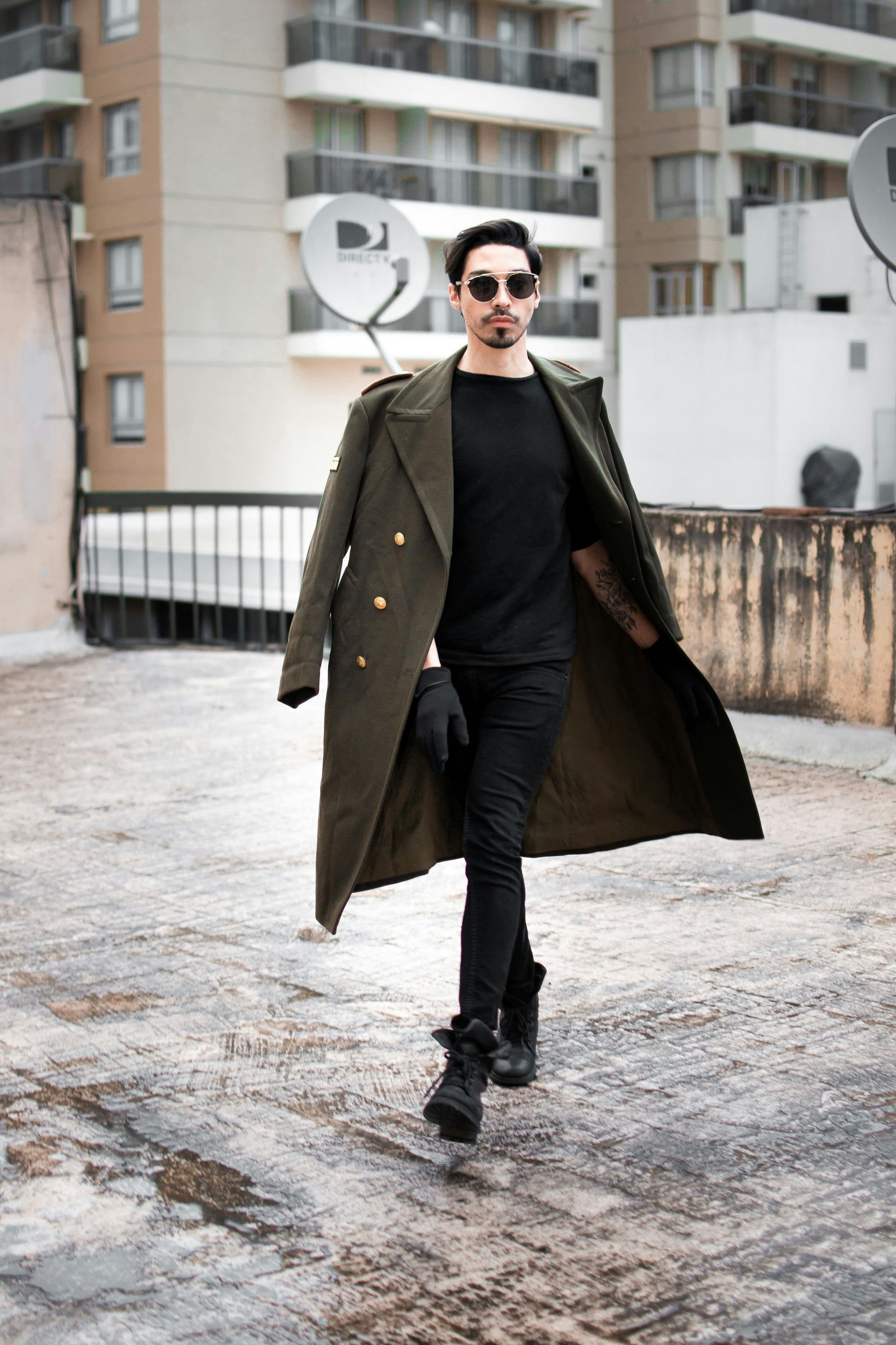 a stylish man walks on a rooftop in the city. he's wearing a long, doublebreasted militarystyle green coat, a black shirt, black pants, and black boots. his sunglasses and the urban background complete the trendy look. the photo is well composed, showing the man from head to toe, and the details of his outfit.