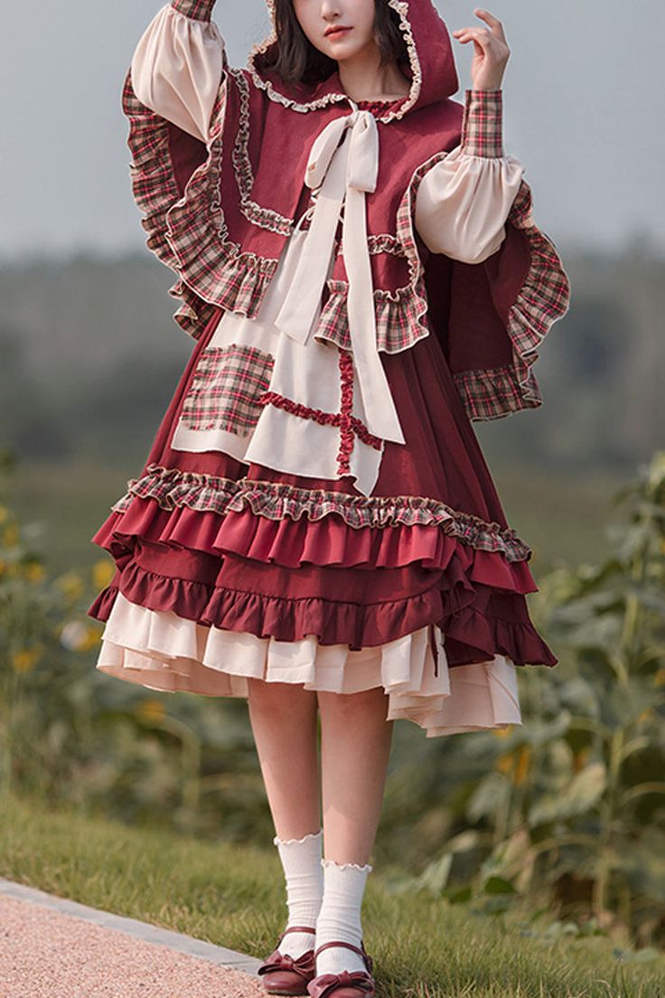 a young woman is modeling a red and cream lolita fashion dress. the dress features a hooded cape, plaid accents, and tiered ruffles. she is standing on a path outdoors, surrounded by greenery, and the photo is taken in natural daylight at eye level.