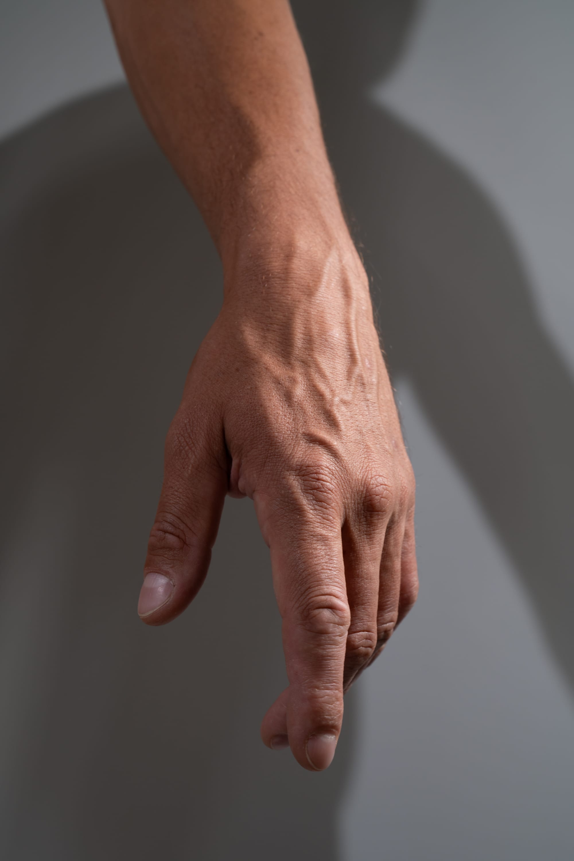 a closeup image of a man's arm and hand.  the lighting is strong, causing the details of his skin and veins to be emphasized. the background is a plain gray wall with a shadow of the arm cast upon it.