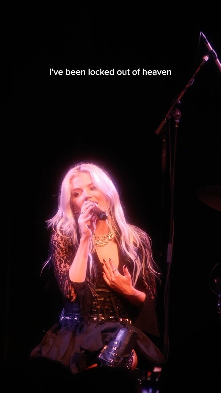 a woman with long blond hair is singing into a microphone on a stage. she is wearing a black outfit with a lace top and a corset. there is a musical instrument behind her. the text "i've been locked out of heaven" is written above her head.