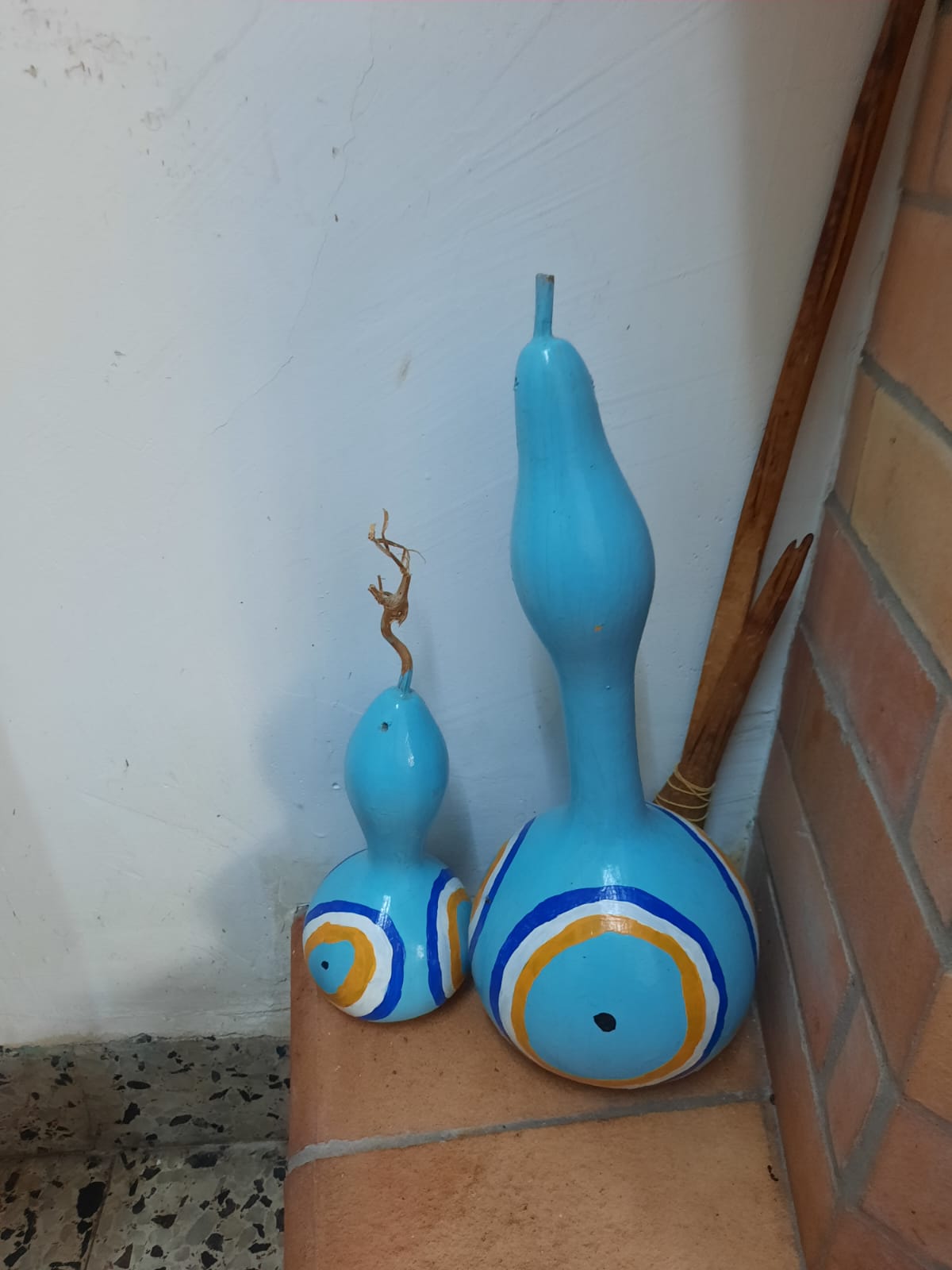 two decorative gourds, painted light blue with concentric bands of white and orange around their bases, stand on a tiled surface against a wall. one gourd is larger with a long, slender neck, while the other is smaller with a twig sticking out of the top. a few sticks lean against the wall behind the larger gourd.