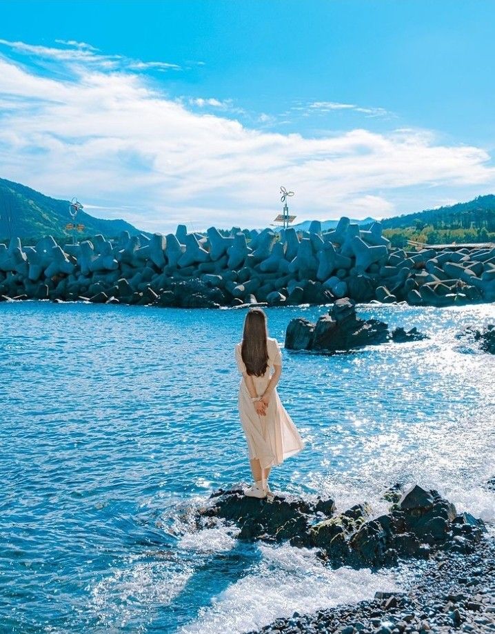a woman in a lightcolored dress stands with her back to the viewer on a rocky outcrop at the edge of a sparkling blue sea. the water is calm except for the waves breaking gently against the rocks. in the background is a tetrapod breakwater, followed by a green mountainous landscape beneath a partly cloudy blue sky.