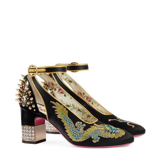 the image showcases a pair of gucci women's highheeled pumps.  the shoes are black satin with elaborate embroidered dragons and floral details. the heels are block style, decorated with crystals and goldcolored studs. the shoes feature a delicate ankle strap closure with a gold buckle. the overall aesthetic is luxurious and combines traditional chinese motifs with modern design elements.  one shoe is slightly angled to show off the detail on the side.