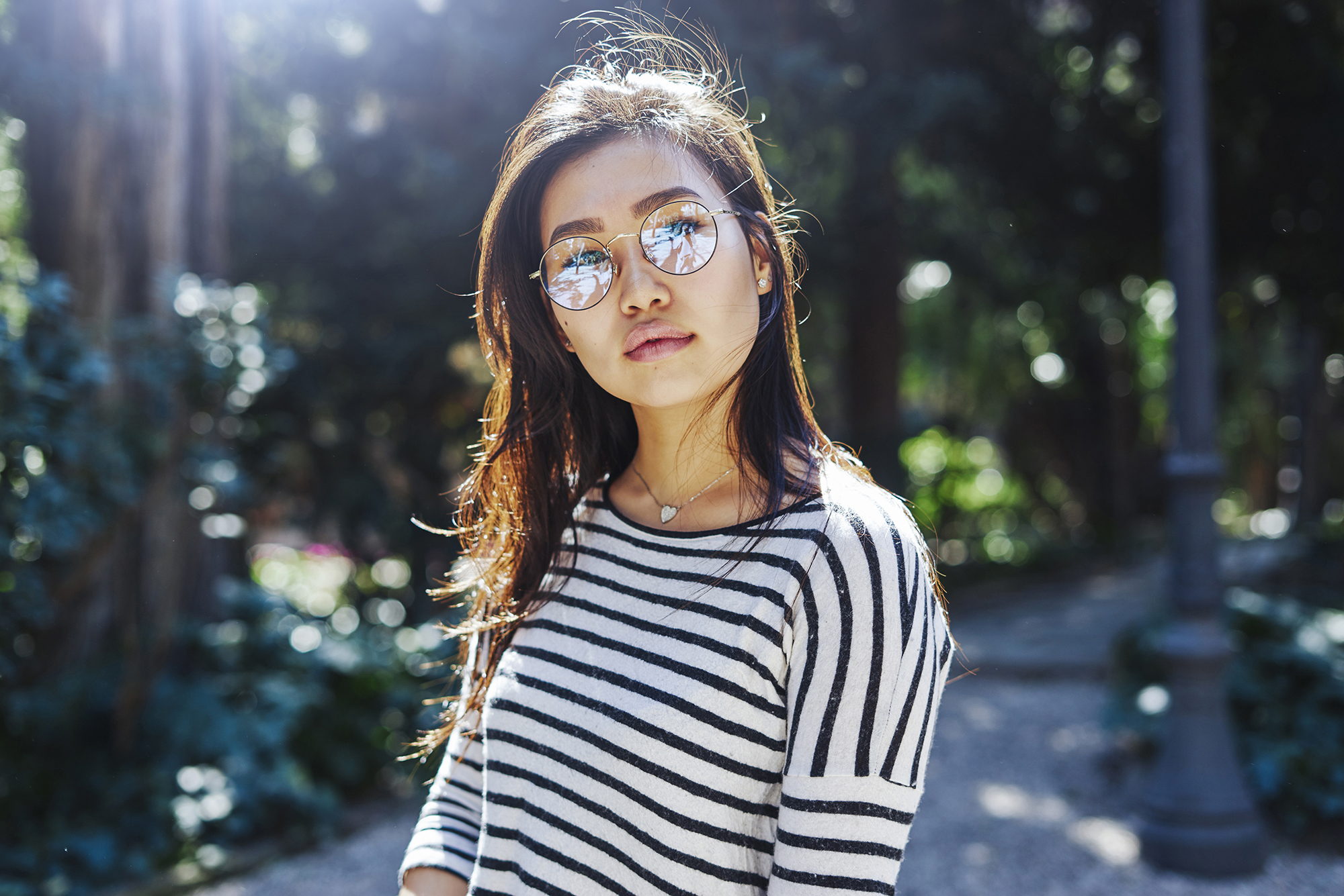 a young woman with long brunette hair is wearing sunglasses and a striped shirt. she is standing outdoors in a park with trees in the background. the sun is shining and the woman is smiling happily.