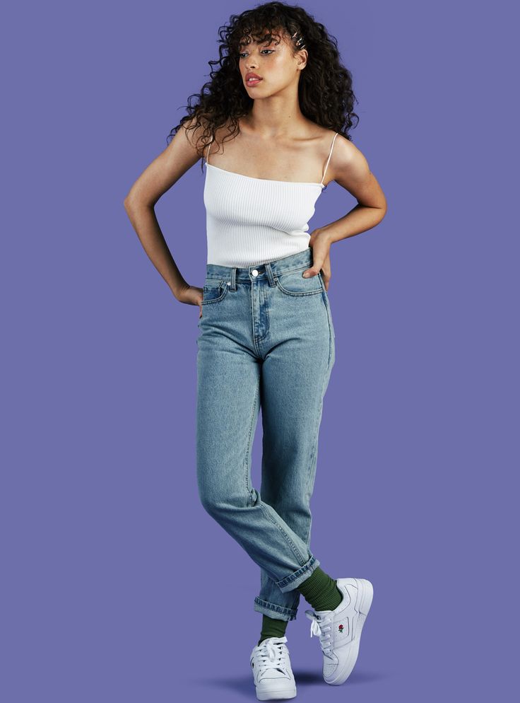 a fullbody shot of a young woman with curly brown hair wearing a white ribbed tank top, light wash straight leg jeans cuffed at the ankles, white sneakers, and olive green socks. she is posing with one leg crossed over the other, hands on her hips, against a purple background.  she has a confident and stylish look.
