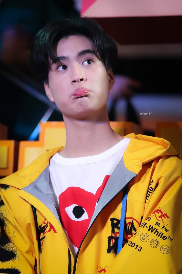 a young asian man with black hair is pictured indoors, wearing a yellow jacket and a white tshirt. he has a slightly pouty expression on his face, with his lips puckered. the background is blurry, but appears to be a store or event space with yellow and orange colors. the lighting is indoor and illuminates the man's face brightly.