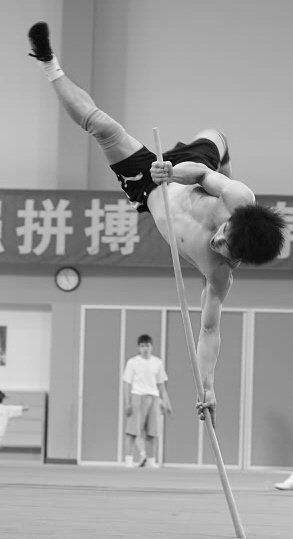 a man in black shorts and a white shirt is doing a gymnastic exercise with a pole. he has his right leg straight up in the air and his left leg bent to the side. he's using his left hand to hold onto the pole for support. his right hand is extended as if he's going to catch something. there is another person in the background. the photo is in black and white.