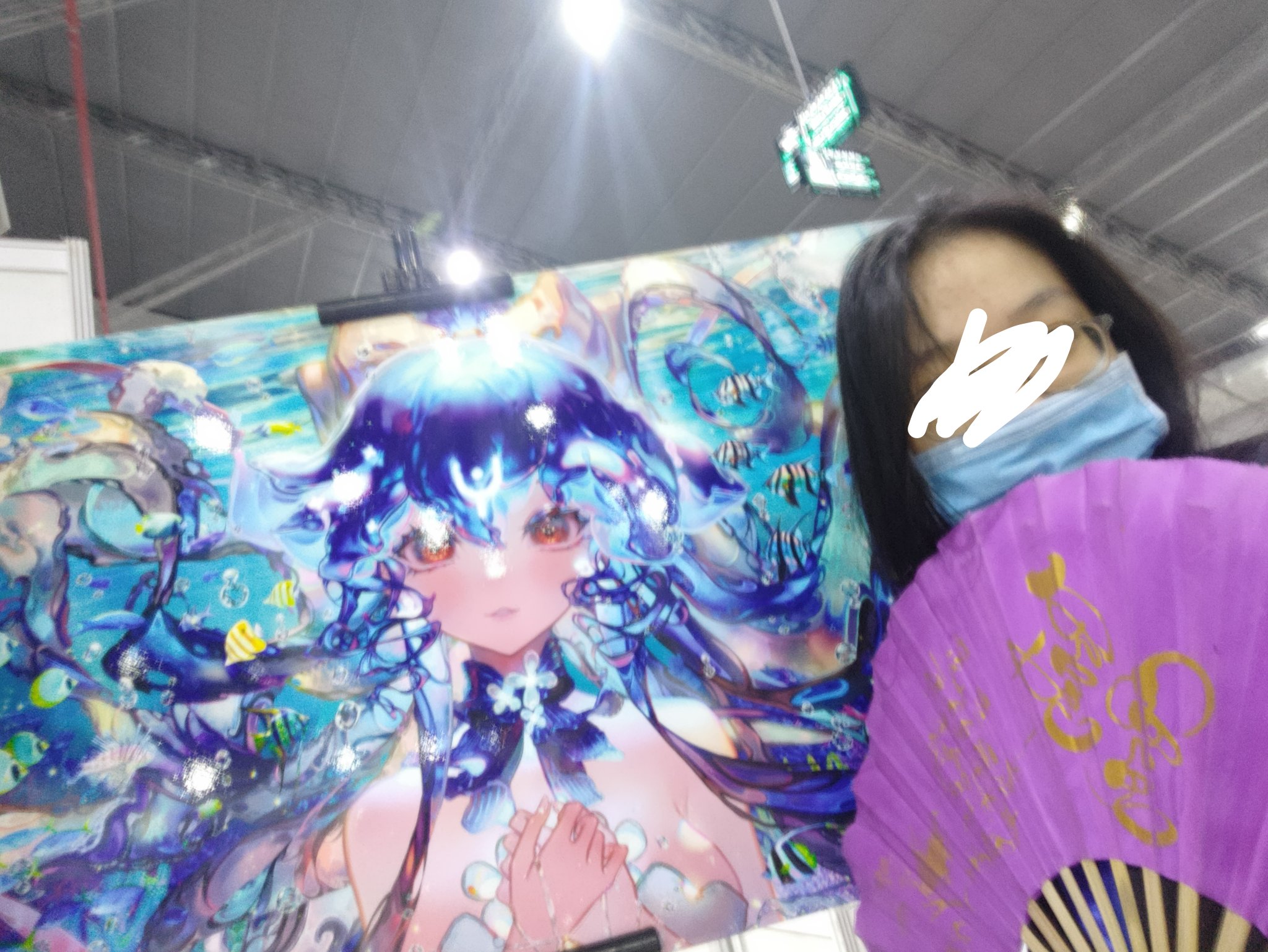 he image shows a person at what appears to be an anime convention, holding a large poster featuring an animestyle illustration of a girl in an underwater scene.  the person is partially obscured, with their face mostly covered by a mask and a purple hand fan. the poster is bright and colorful, depicting a young woman with long blue hair surrounded by fish and other marine life. the overall atmosphere is vibrant and related to anime/manga culture.