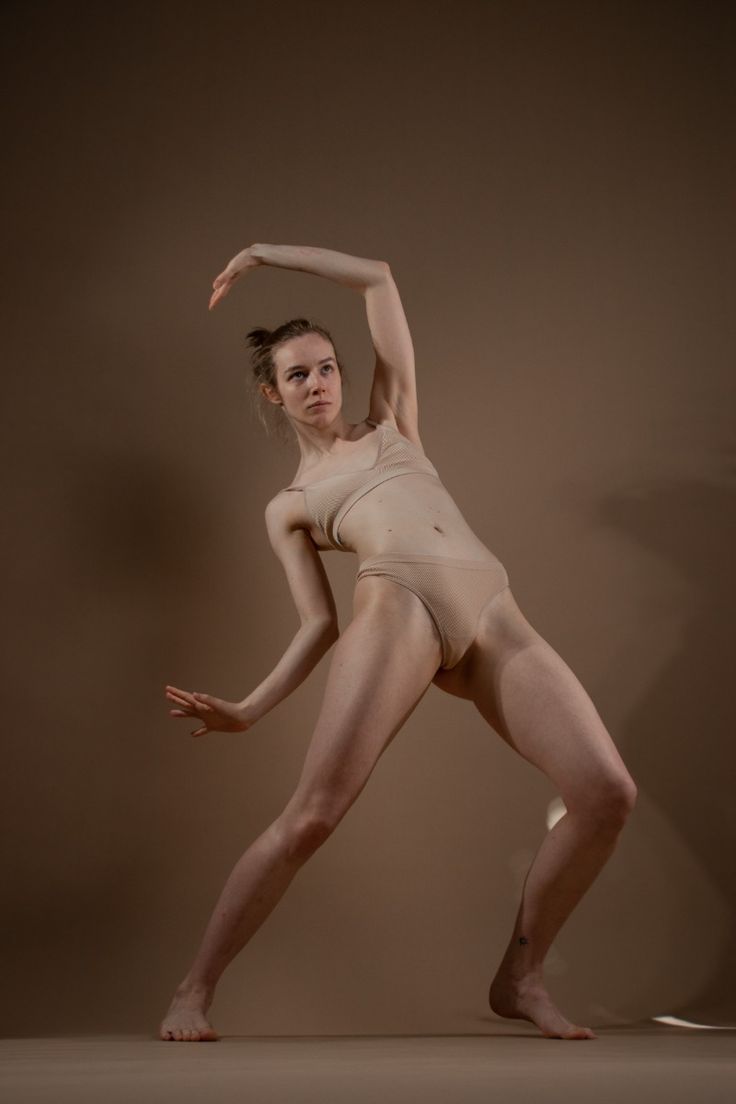 a woman in nude underwear is in a dance pose in a studio against a brown background. one arm is raised and bent, the other is extended to the side. her legs are apart.