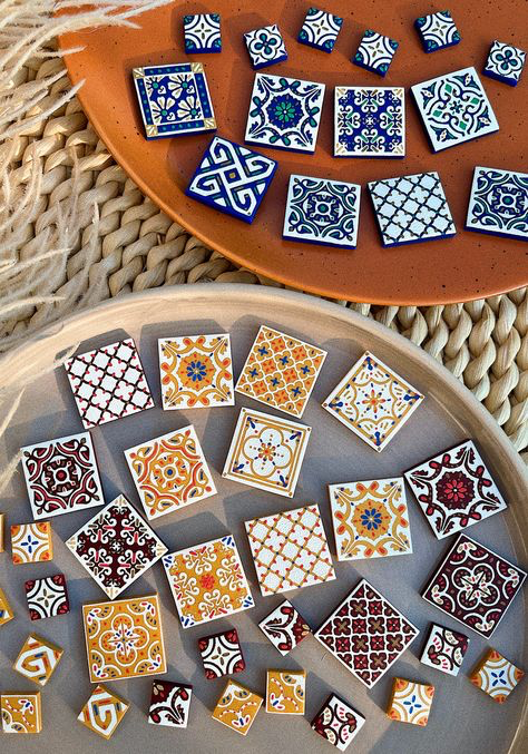 the image shows a collection of small, square ceramic tile earrings arranged on two circular trays.  the trays rest on a woven surface.  there are two distinct styles of tiles: one set features blue, white, and gold designs, while the other set is primarily in shades of orange, yellow, red and brown with different patterns. the earrings are meticulously crafted and showcase various geometric and floral patterns reminiscent of traditional styles. the overall aesthetic is bohemian and suggests handmade or artisan jewelry. the flatlay photography style emphasizes the vibrant colors and intricate details of the earrings and the warm tones of the surrounding setting.