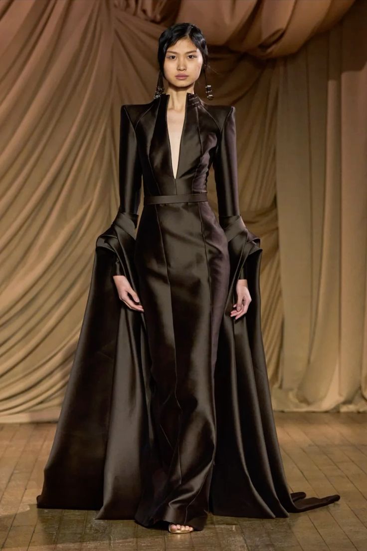 a female model is walking on a runway in a black gown with a long cape. the gown is made of satin and the cape is made of silk. the model is wearing a pair of earrings and has her hair pulled back. the background is a beige backdrop with draping and a wooden floor.  this is a photo from a highfashion runway show.