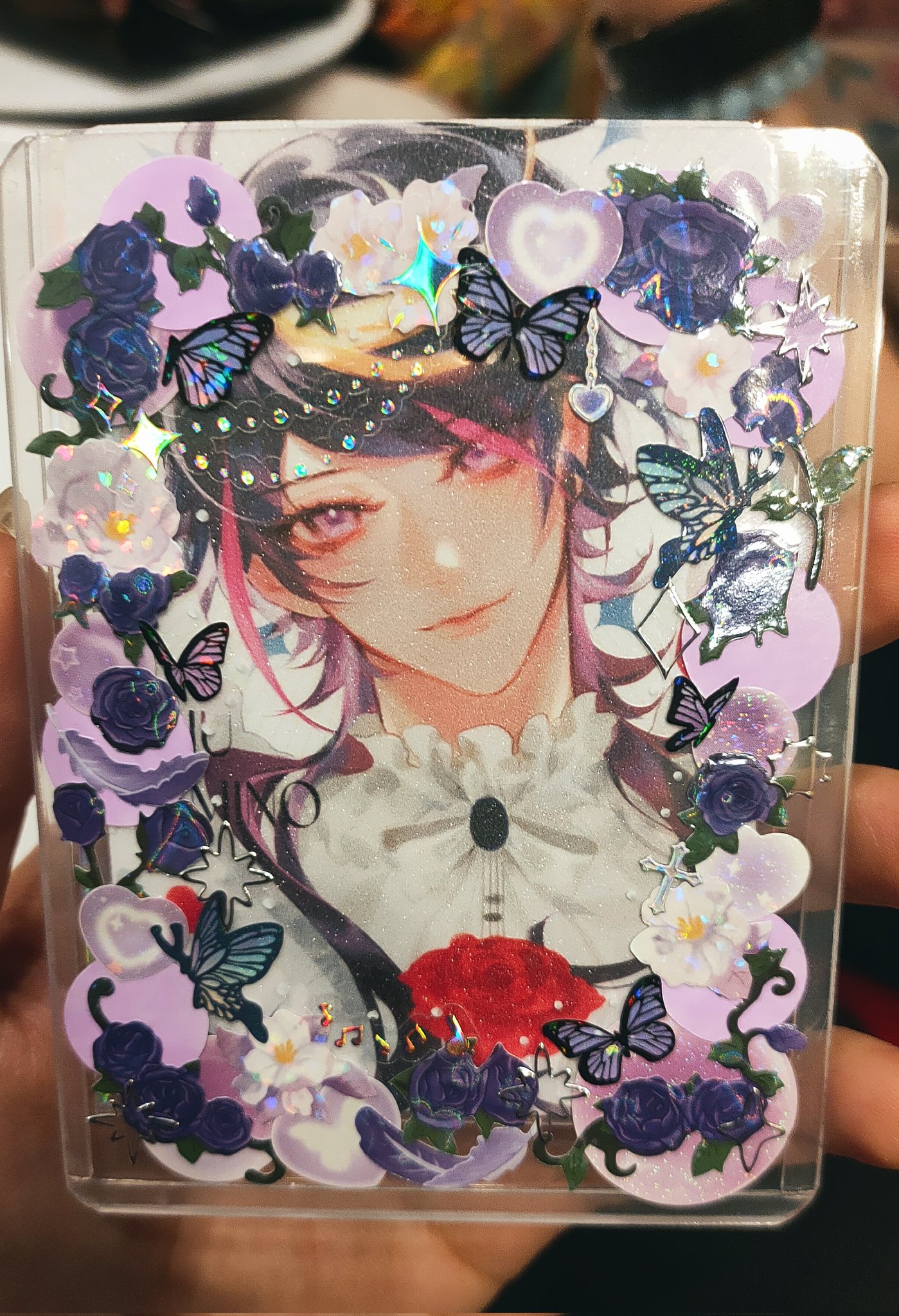closeup view of a handmade resin art piece.  it's a clear plastic case or card protector containing a printed image of an animestyle boy with purple hair. the image is surrounded by various embellishments including purple and white flowers, butterflies, glitter, and iridescent accents. the overall aesthetic is cute and kawaii.  the resin is clear and glossy, showcasing the details of the artwork and decorations.  a hand is holding the resin piece in the bottom right corner of the picture.