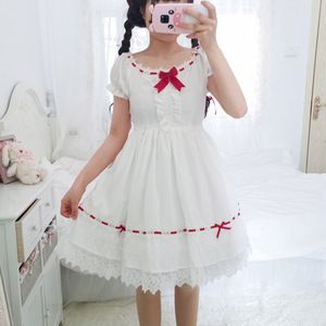 a young woman is posing in a white lolita dress. she's standing in a bedroom, facing the camera. the dress has red ribbon detail and lace trim.  the dress is short and puffy, with a square neckline and puffed sleeves.  the overall aesthetic is sweet and girly.
