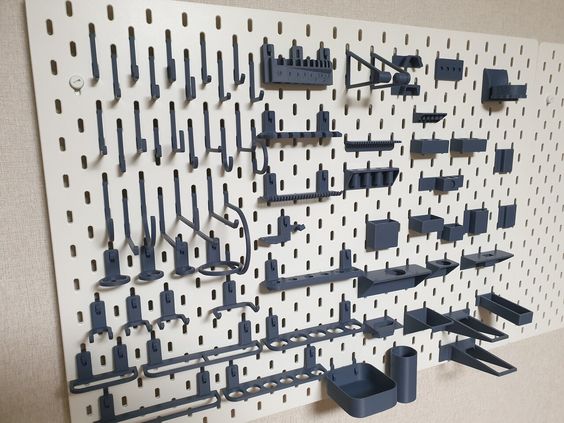 a white pegboard wall with a variety of 3d printed pegboard hooks and accessories. the hooks are in a variety of shapes and sizes, and they are arranged in a pattern on the wall. the accessories include shelves, baskets, and a variety of other storage solutions. the pegboard is a great way to organize a workspace or a garage.