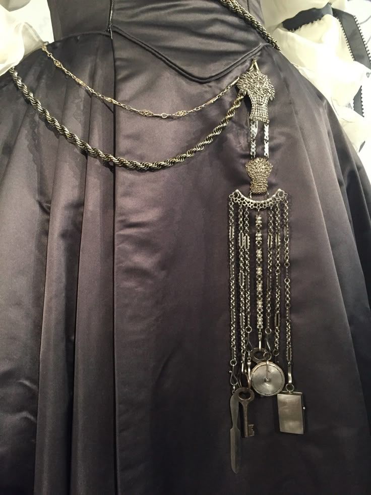 a closeup shot shows a black satin dress or skirt with a silver chain beltlike accessory. the chain hangs with various trinkets, including a key, a watch, and other metallic ornaments. the chain has multiple strands, and the trinkets dangle from the bottom. the dress's fabric has a smooth and reflective quality.