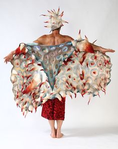 a person is wearing a very large, elaborate costume made of felt, feathers, and other materials. the costume is very intricate and detailed, and it appears to be inspired by nature, particularly the ocean or sea life. the costume is very colorful and abstract, and it creates a sense of movement and energy. the person is standing in a studio, and their back is to the camera. the costume is the focus of the image, and it is clearly a work of art. the person appears to be a performer or artist, and the costume is a testament to their creativity and imagination.
