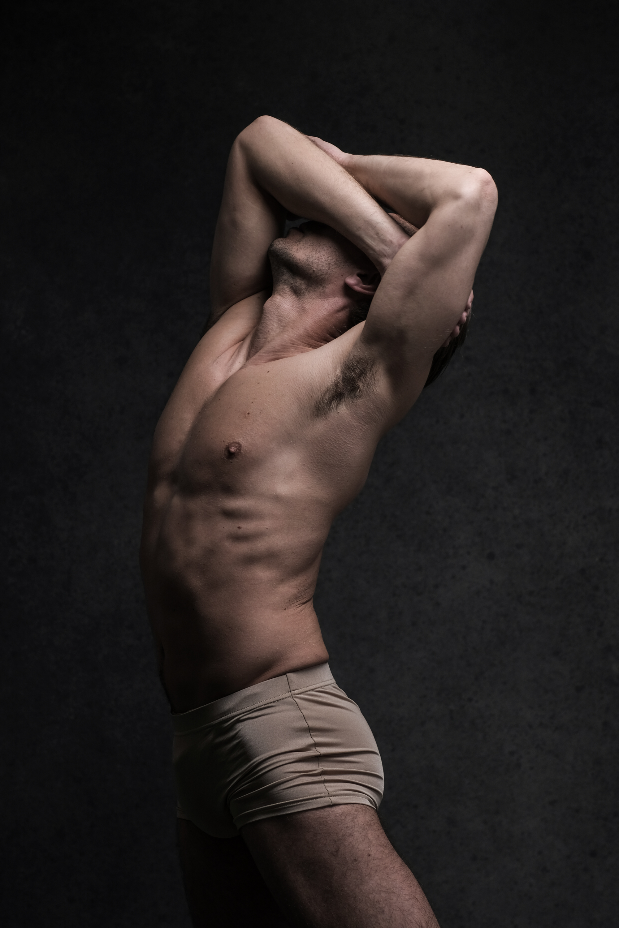the image showcases a muscular male figure, likely a dancer, in a dynamic pose against a dark studio backdrop. he is wearing only underwear, highlighting his toned physique. the studio lighting is dramatic, casting shadows that accentuate his musculature and the contours of his body.  the perspective is a side view, capturing the stretch and extension of his limbs.