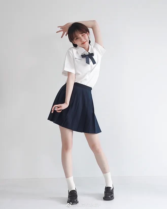 a young woman in a school uniform is posing in front of a white background. she is wearing a white shirt with a navy blue bow, a navy blue pleated skirt, white socks, and black loafers. she has short brown hair and is smiling slightly. she is holding one arm above her head.