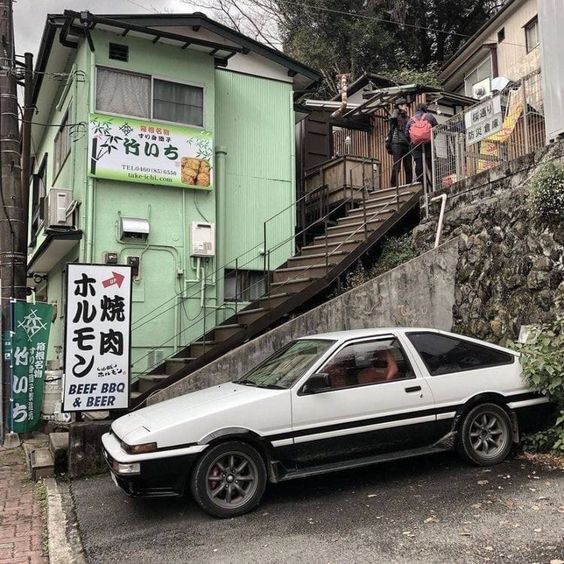 a white and black toyota ae86 sprinter trueno is parked on the street in front of a green building with japanese signage. two people are walking up a set of stairs to the right of the building. the scene appears to be in japan and has a nostalgic, street photography vibe.