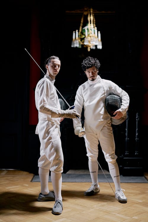 two male fencers stand in a dimly lit room with a chandelier in the background.  they are dressed in formal white fencing uniforms, holding foils and wearing masks. the photo is professionally shot with a focus on the athletes and their attire. the overall mood is elegant and classic.