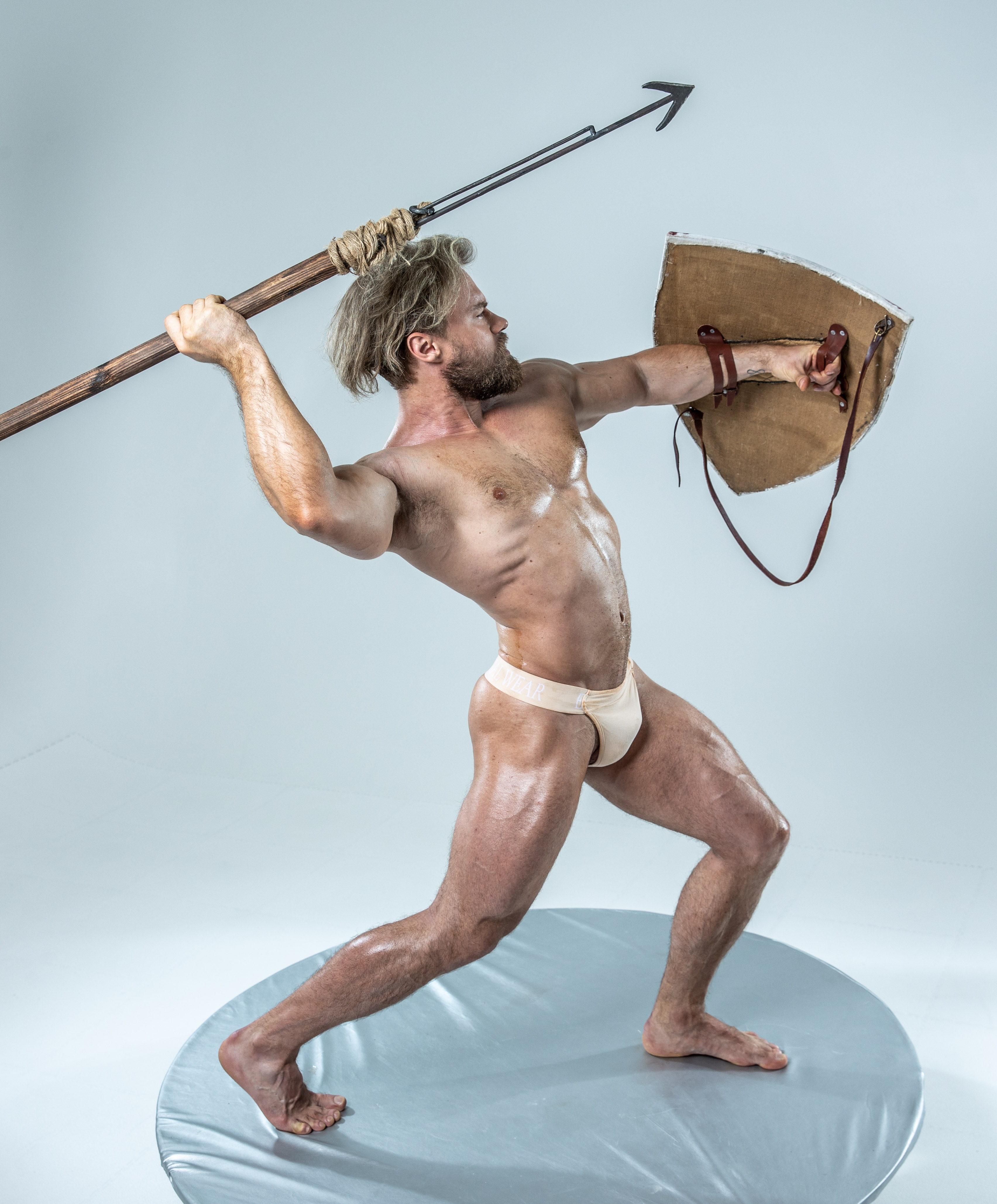 a muscular man in a warrior pose, wielding a spear and shield against a white studio backdrop. the lighting is soft, highlighting his physique and the texture of his weapons. his expression is fierce, capturing the essence of battle.
